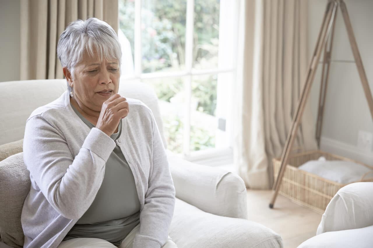Older woman coughing while sitting in her living room at home.