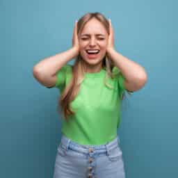 A young woman in a casual outfit covered her ears from a loud sound on a blue isolated background